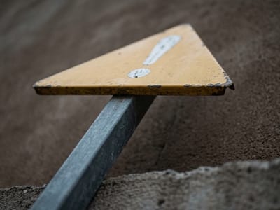 Yellow triangular sign on a leaning metal post against bare brown ground