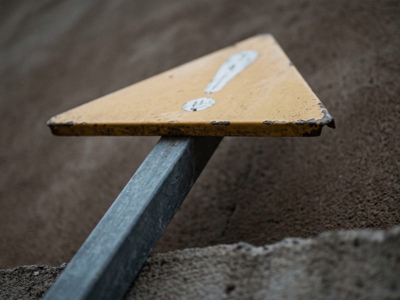 Yellow triangular sign on a leaning metal post against bare brown ground