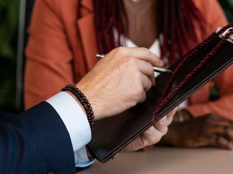 Hand with a beaded bracelet holding a pen over a leather folder during a meeting